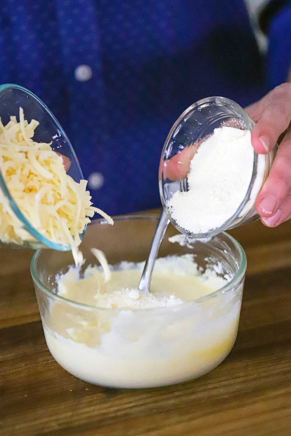 A person using two hands to dump shredded mozzarella in a small bowl in one hand and grated parmesan from a bowl in the other hand into a bowl of ricotta. 