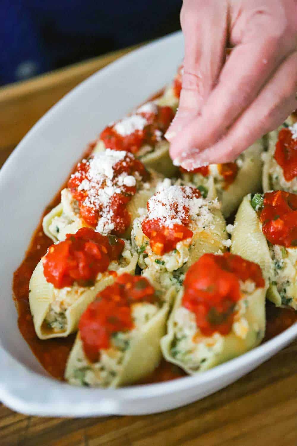 A hand sprinkling grated Parmesan cheese over the tops of uncooked stuffed shells in a white baking dish. 