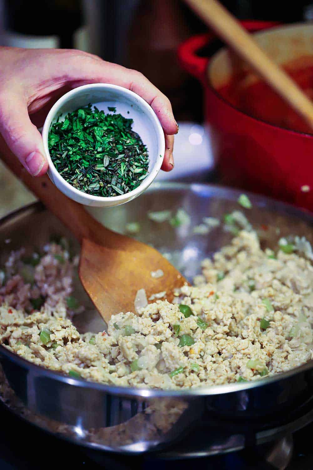 A hand dumping freshly chopped herbs from a small bowl into a large skillet filled with cooked chicken sausage. 
