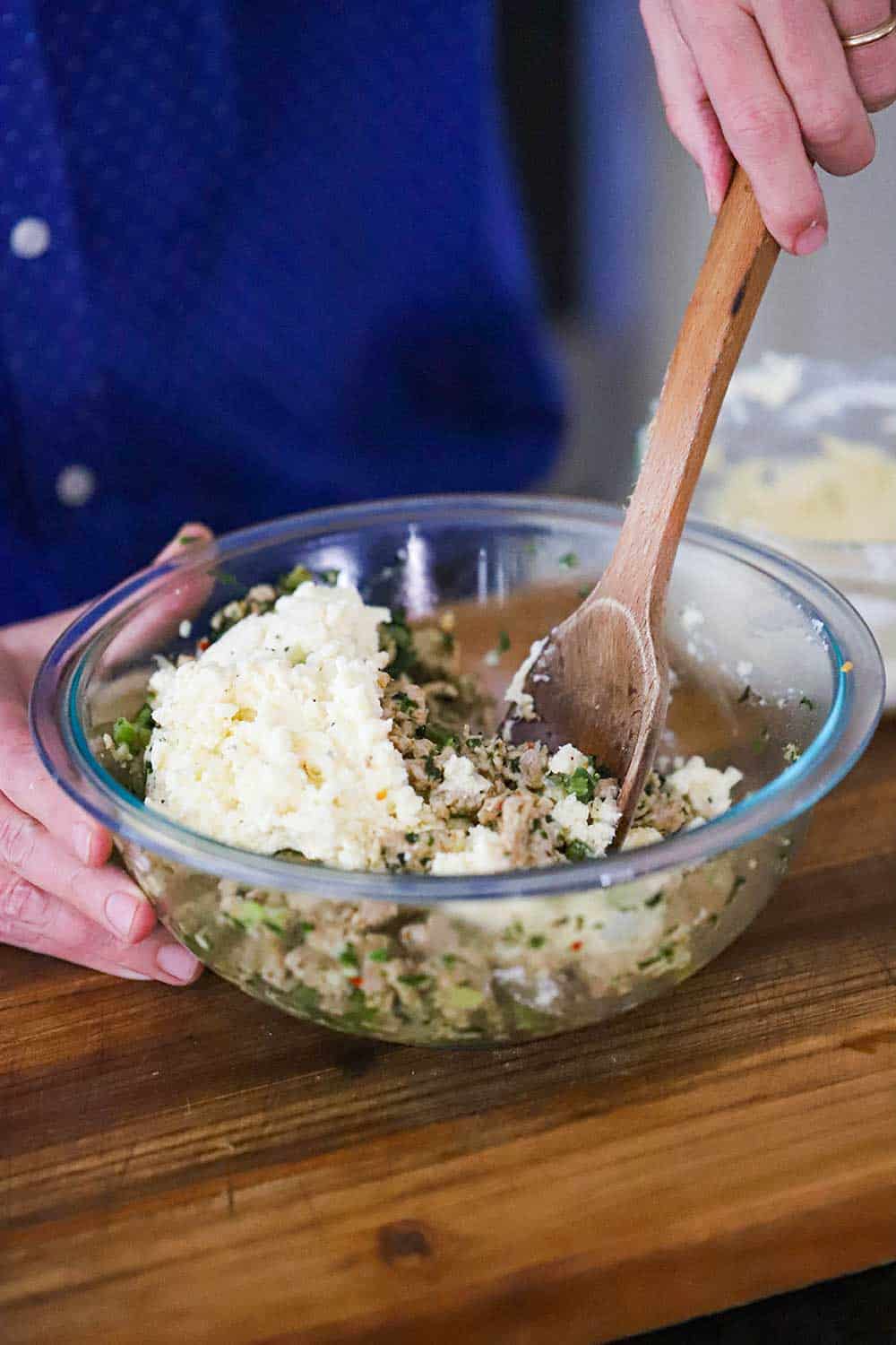 A person using a large wooden spoon to stir together a cooked chicken mixture in a glass bowl with a ricotta and mozzarella mixture. 