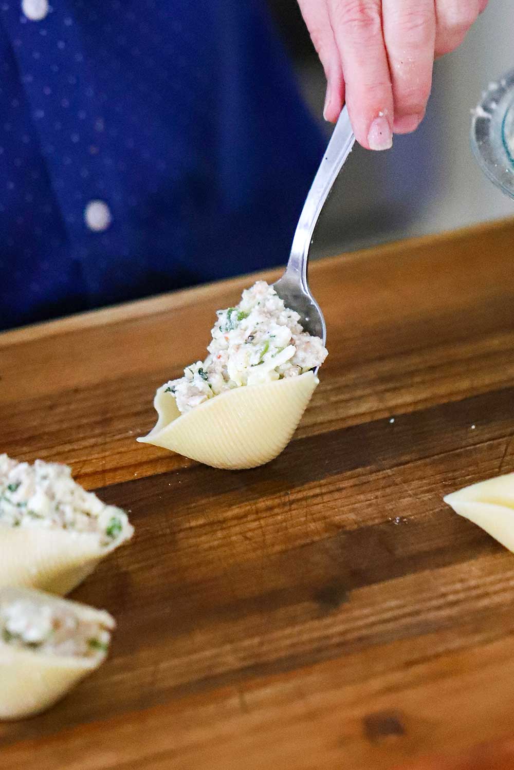 A person using a large spoon to add a cheese and cooked sausage into a jumbo shell. 