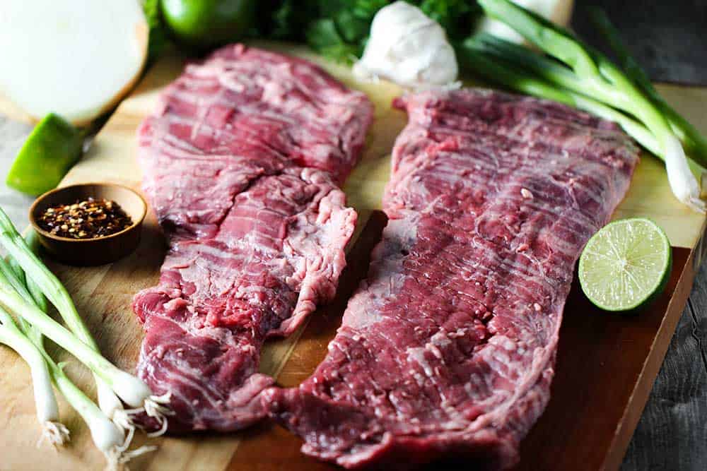 Two large cuts of skirt steak on a butting board next to cut limes and scallions. 