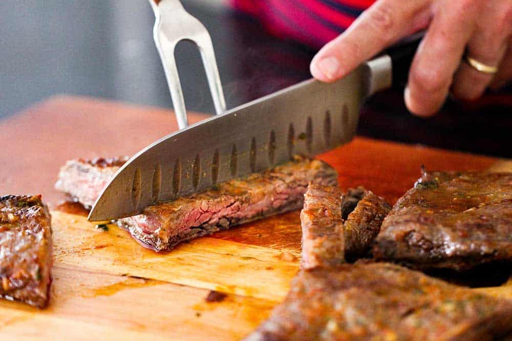 A large knife cutting a cooked skirt steak against the grain on a cutting board. 