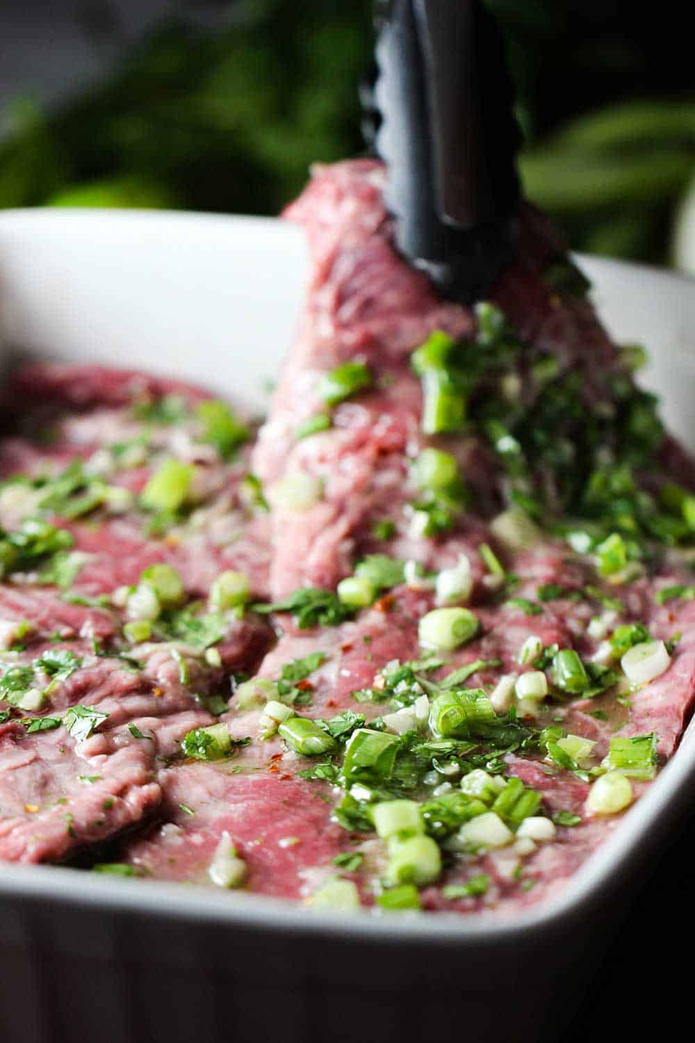 A pair of tongs lifting a cut of skirt steak up that has been marinating in a white dish. 