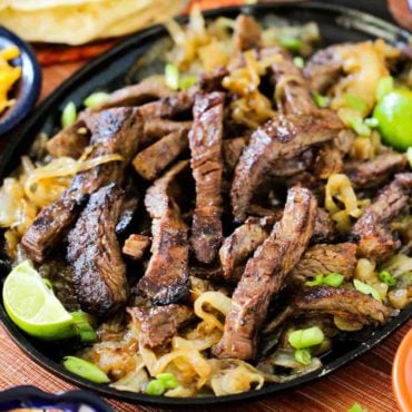 Authentic steak fajitas on a large fajita pan with sliced limes and scallions nearby.