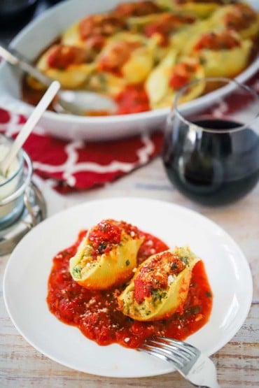 Two Italian stuffed shells on a plate with a layer of marinara sauce on the bottom all sitting next to a glass of red wine.
