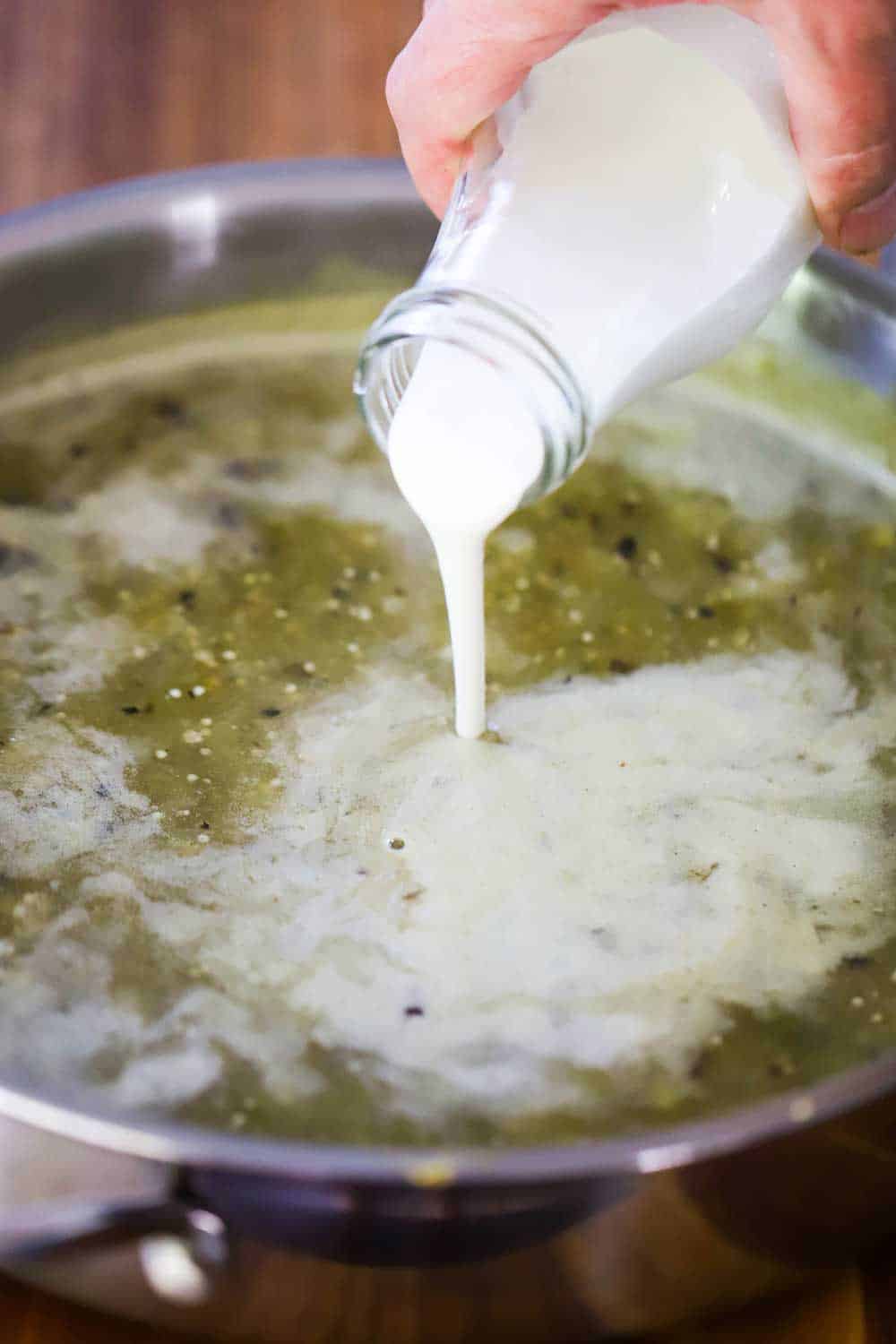 A hand pouring Mexican crema from a bottle into a skillet filled with hot verdi sauce. 