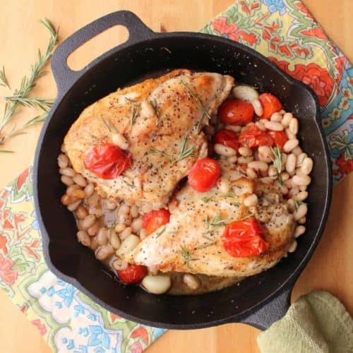 A large cast-iron skillSkillet-roasted chicken with tomatoes, white beans and wine sitting in a large cast-iron skillet next to festive napkins.