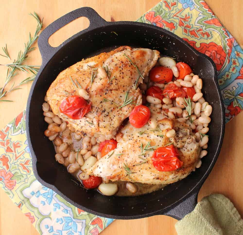 A large cast-iron skillSkillet-roasted chicken with tomatoes, white beans and wine sitting in a large cast-iron skillet next to festive napkins.