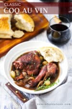 A white plate holding Coq au Vin with a piece of a loaf of bread next to it, and a stemless glass of red wine and a loaf of bread nearby.