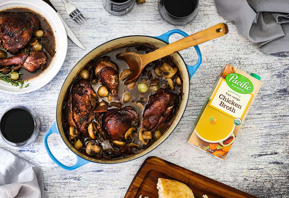 An overhead view looking down at a large Dutch oven filled with Coq au Vin next to a plate of the dish and next to a box of chicken broth.