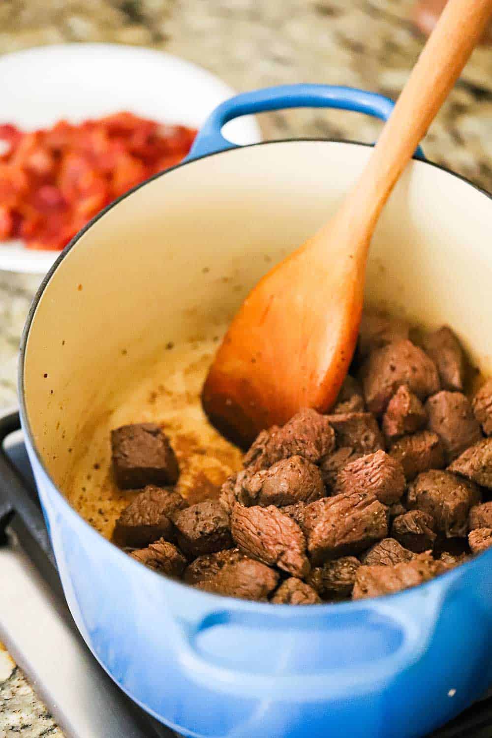 A large blue Dutch oven filled with pieces of chuck beef roast that have been seared with a wooden spoon in the pot. 