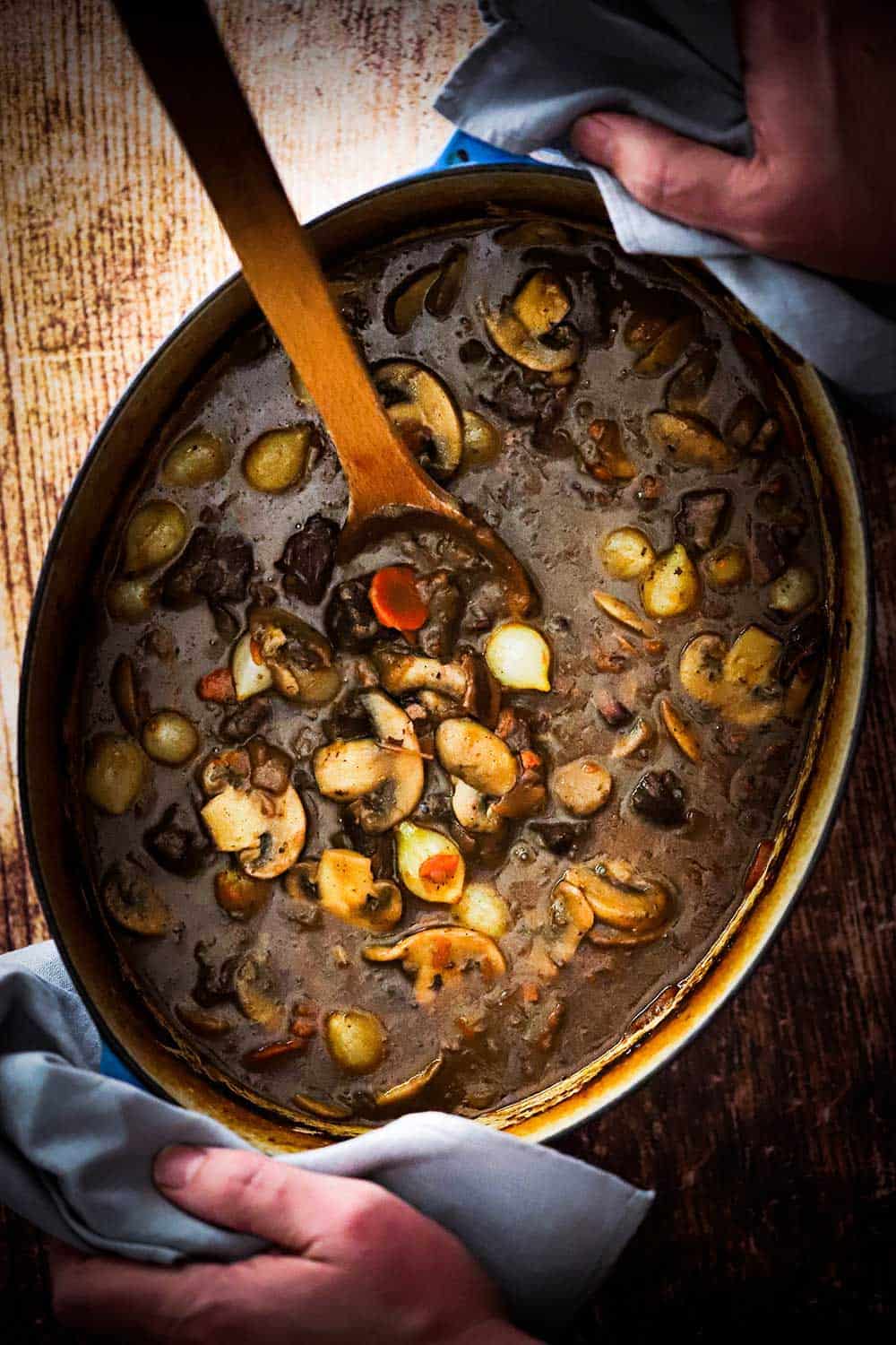 A large oval Dutch oven filled with beef bourguignon being held by two hands. 