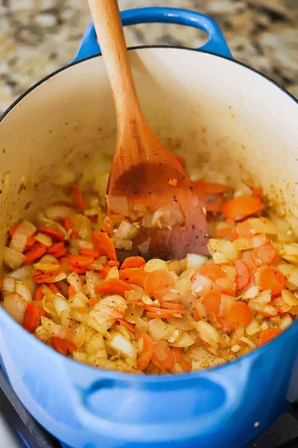 A blue Dutch oven filled with sautéd onions and carrots and a wooden spoon in the middle of it all. 