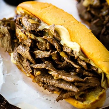 A large Philly cheesesteak loaded with sliced cooked ribeye and melted Provolone cheese sitting on white butcher paper.