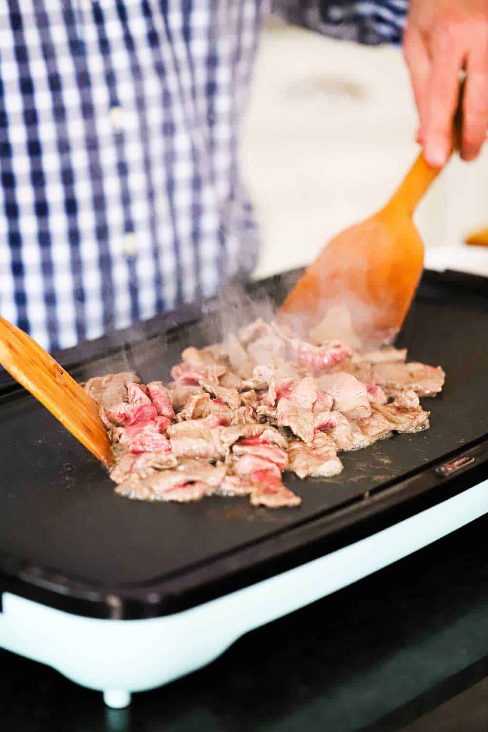 A person using two wooden spatulas to cook and stir thin slices of ribeye steak on a large indoor griddle. 