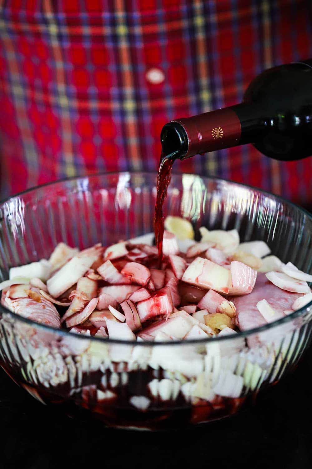 A person in a red shirt pouring a bottle of red wine into a large glass bowl filled with chicken pieces and chopped onion. 