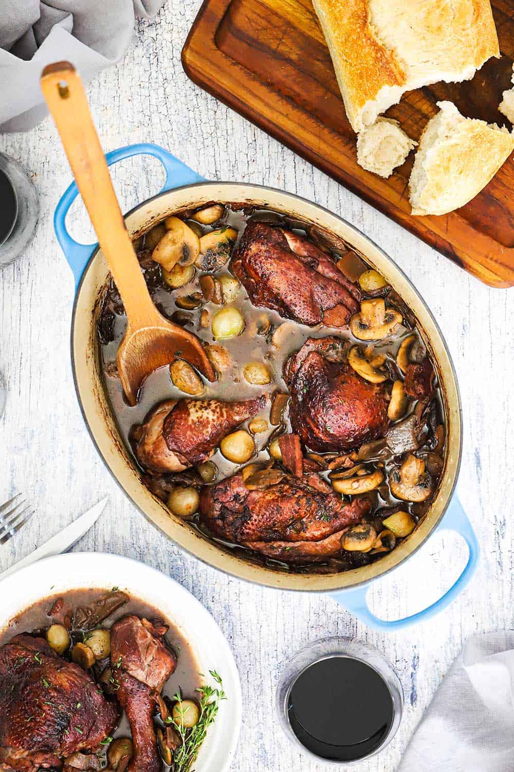 An overhead view of a large oval-shaped Dutch oven filled with Coq au Vin with a wooden spoon in and next to a cutting board topped with bread and next to a glass of red wine. 