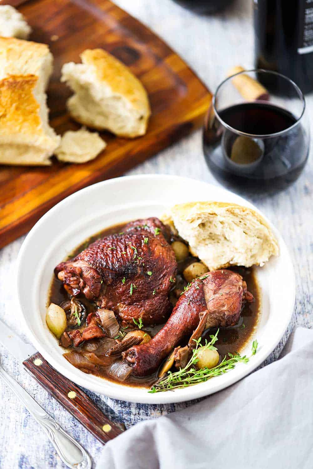 A white plate holding Coq au Vin with a piece of a loaf of bread next to it, and a stemless glass of red wine and a loaf of bread nearby. 