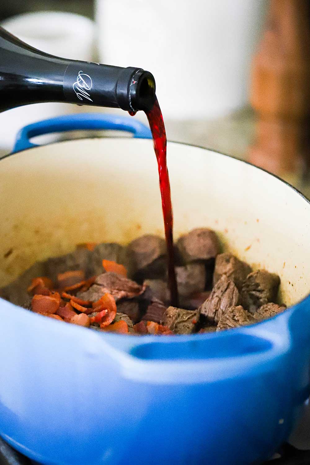 A bottle of red wine being poured into a Dutch oven filled with seared beef and vegetables. 