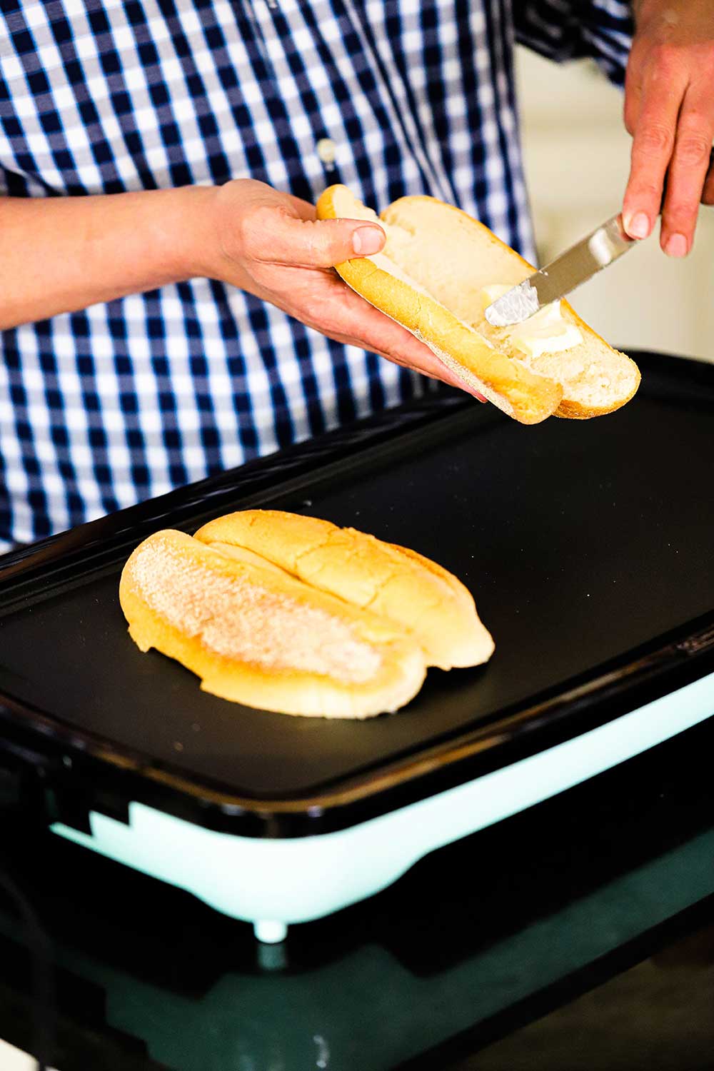 A person smearing softened butter onto the inside of a cut open hoagie over a griddle with another hoagies being toasted, cut-side down. 