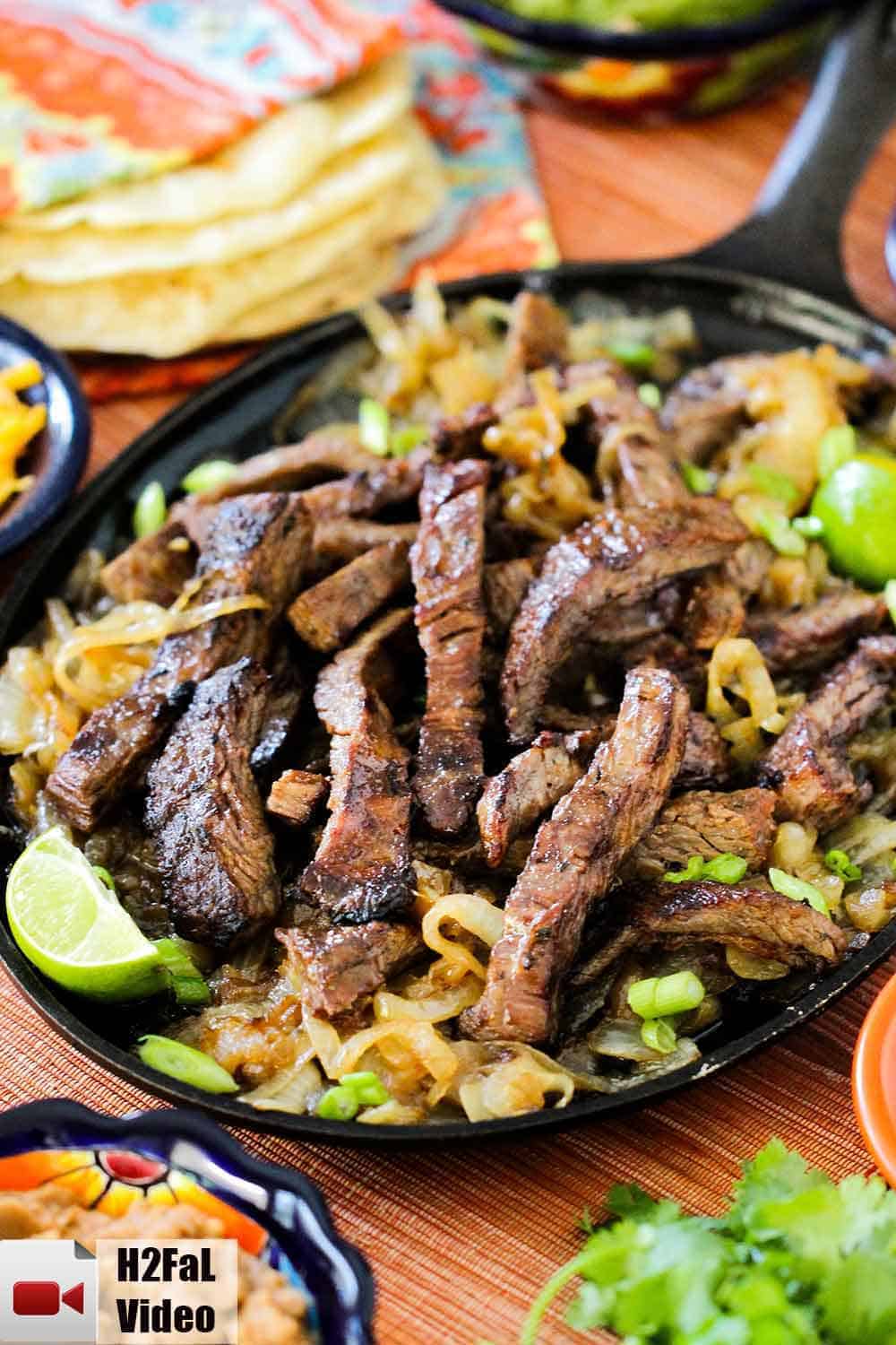 A cast iron fajita skillet of steak fajitas with limes and green onions nearby.