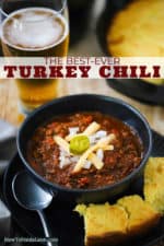 A dark bowl filled with turkey chili topped with shredded cheese and a sliced jalapeno all sitting next to a tall glass of beer.