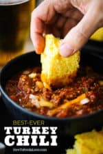 A hand dipping a slice of homemade cornbread into a bowl of turkey chili.