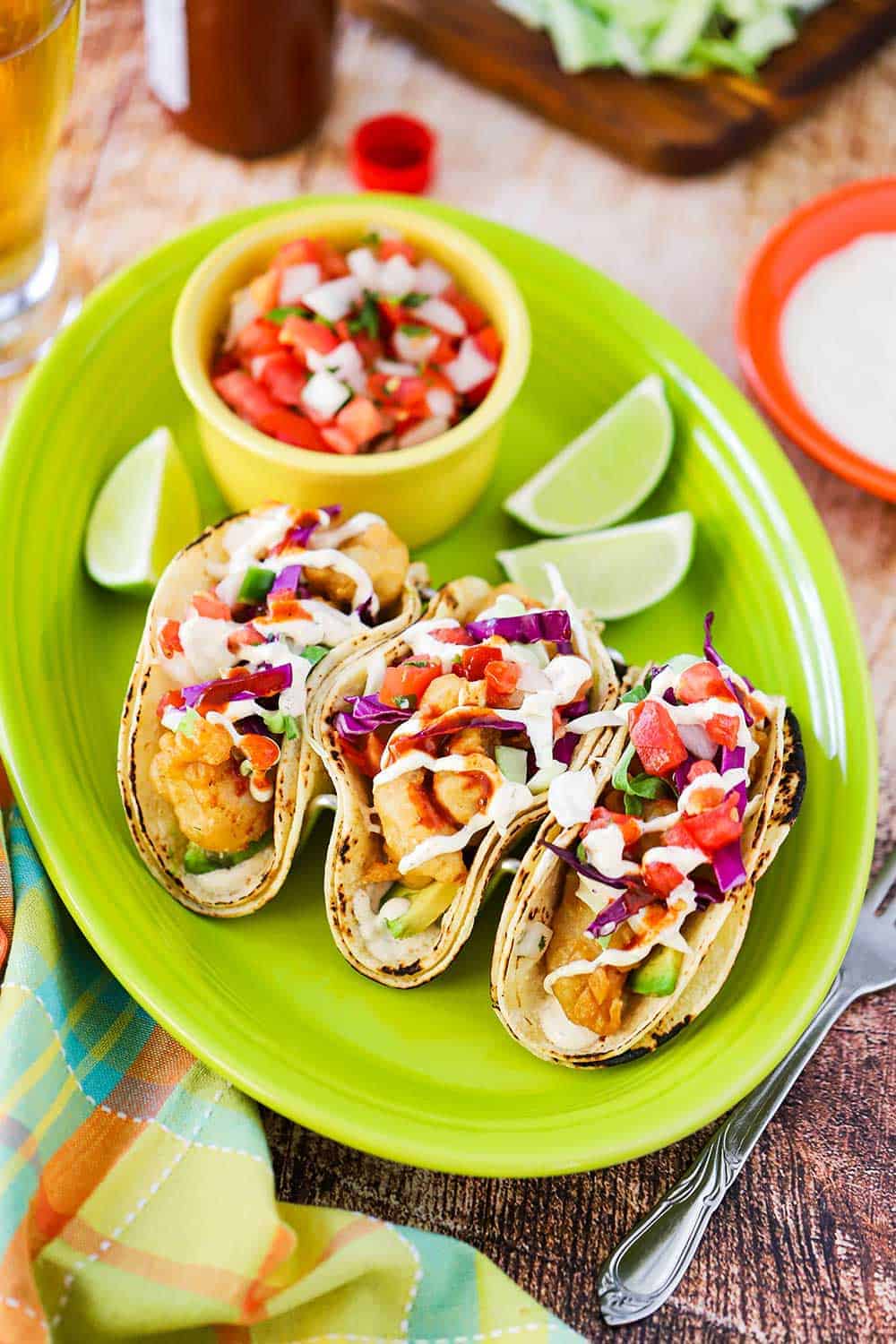 An overhead view of three fish tacos sitting on a green platter next to lime wedges and a bowl of pico de gallo.