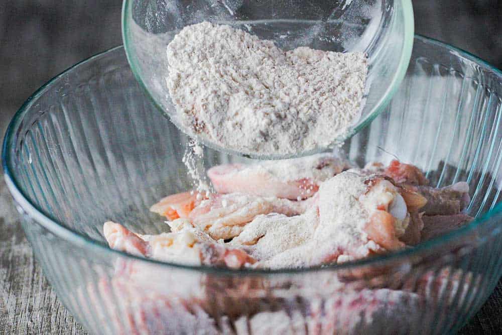 A hand sprinkling seasoned flour onto chicken wings in a glass bowl. 