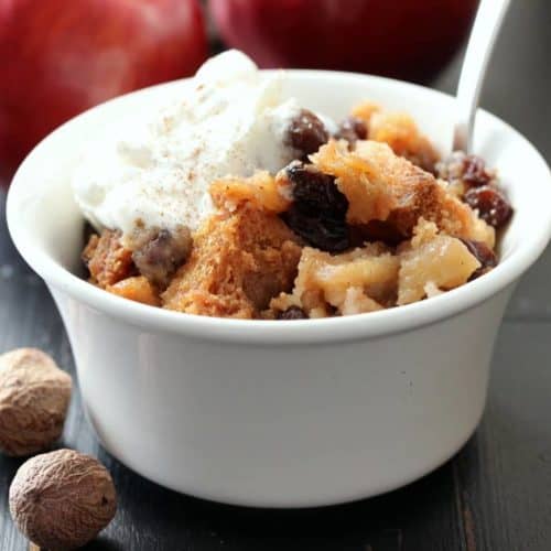 A white bowl with apple brown better with a silver spoon in next to whole nutmegs.