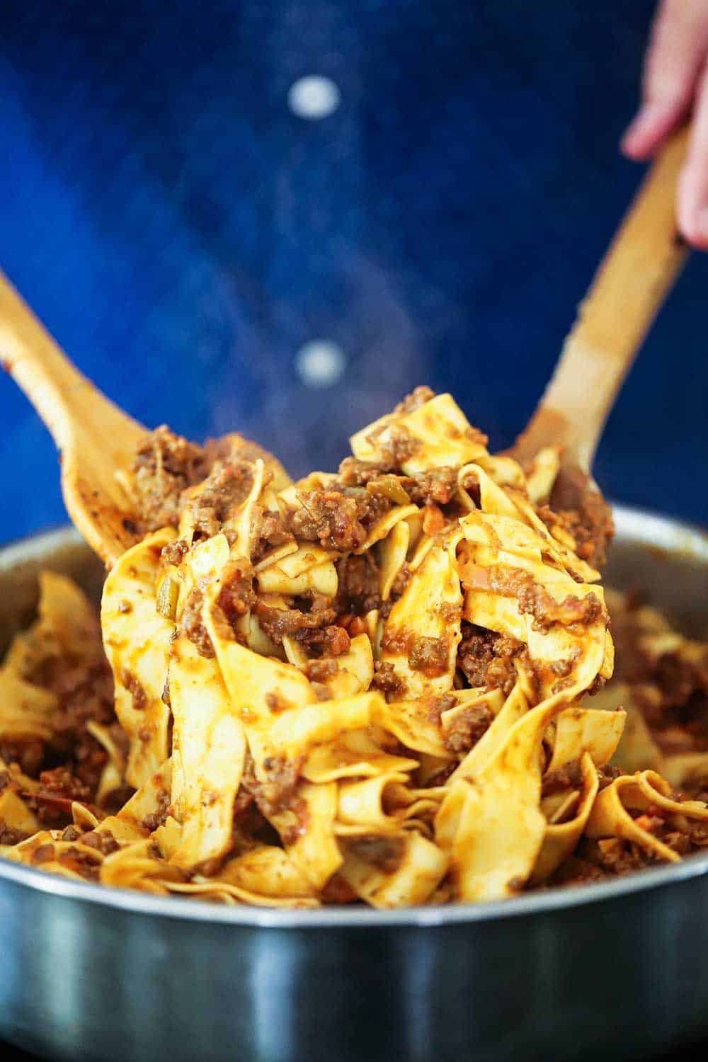 Two hands holding two wooden spoons tossing pappardelle bolognese in a large silver skillet. 