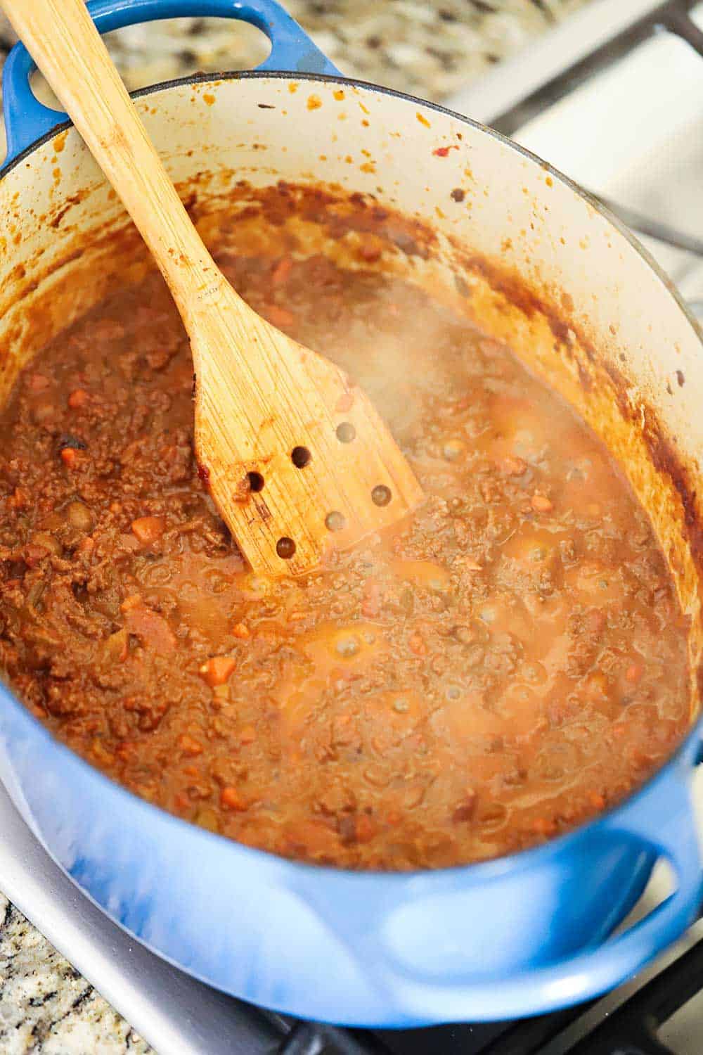 A large blue Dutch oven sitting on a stovetop filled with meat bolognese sauce with a wooden spoon in it. 