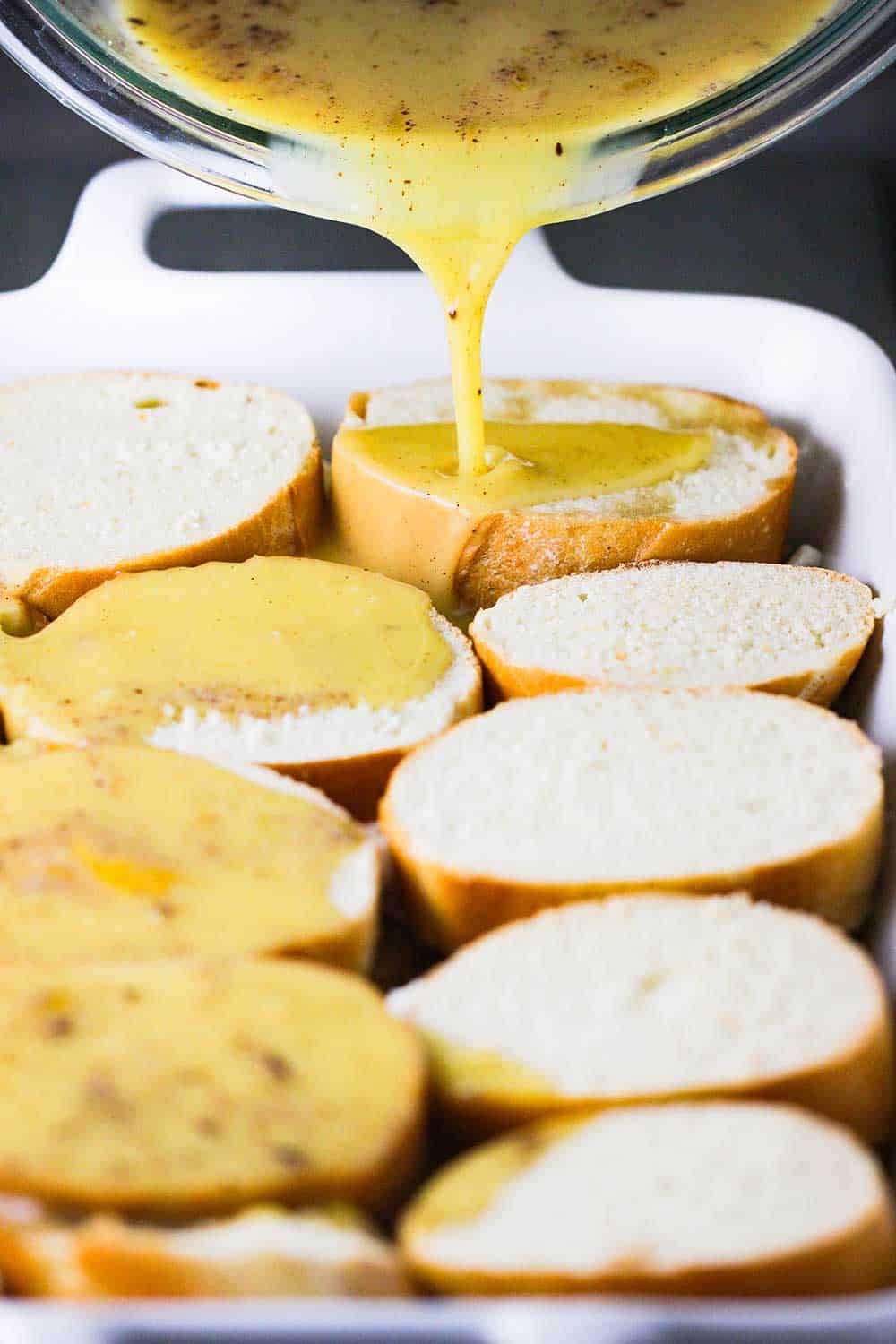 A bowl pouring an egg mixture over a layer of French bread slices in a large white baking dish. 