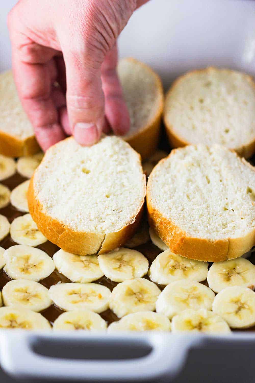 A hand placing slices of French bread over a layer of slice bananas and caramel sauce in a white baking dish. 