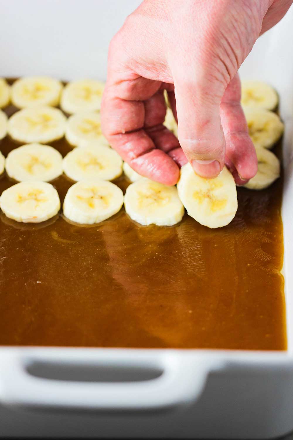 A hand placing sliced bananas on top of a layer of caramel sauce in a white baking dish. 