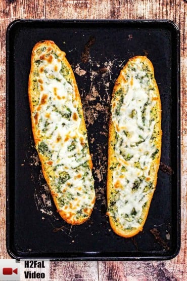 Two loaves of cheesy garlic bread on a scratched black baking pan.