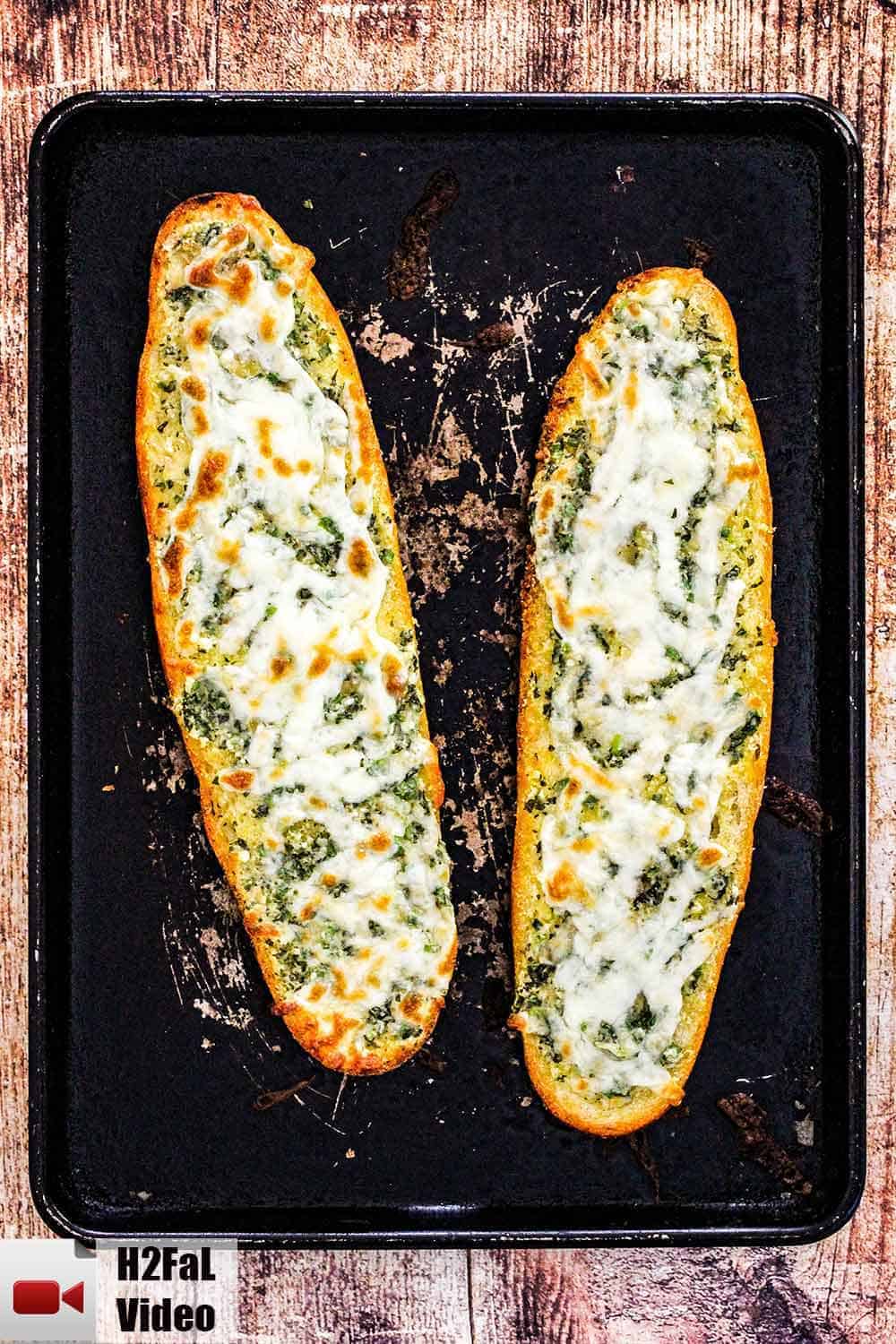 Two loaves of cheesy garlic bread on a scratched black baking pan. 