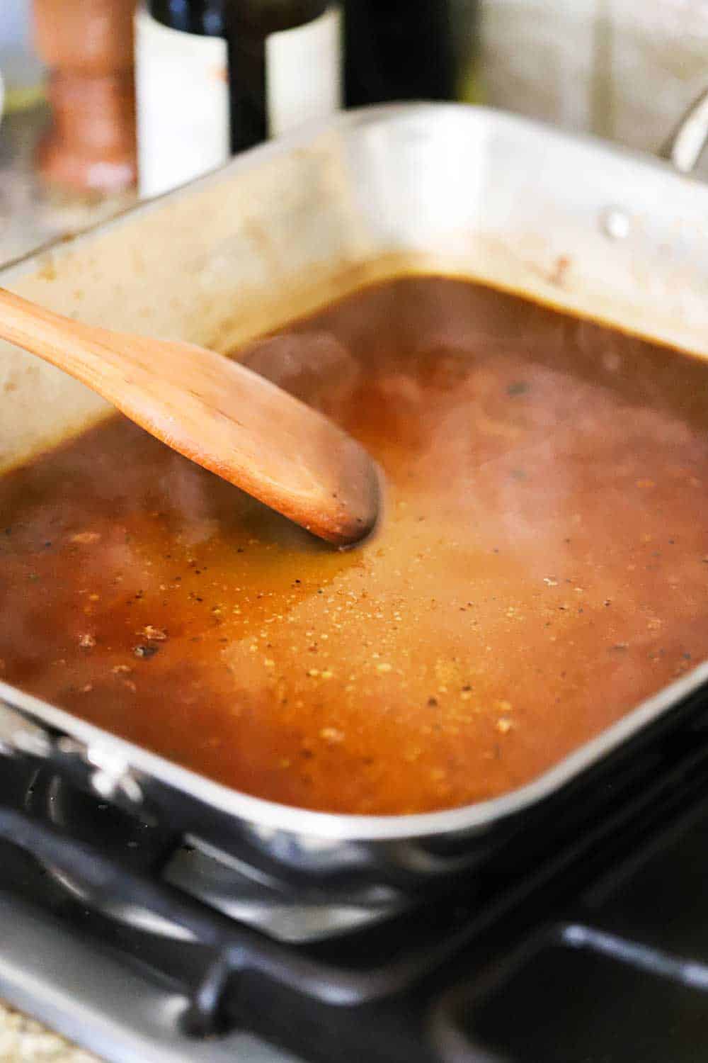 A large silver roasting pan filled with roast chicken drippings and broth being stirred with a wooden spatula while simmering. 