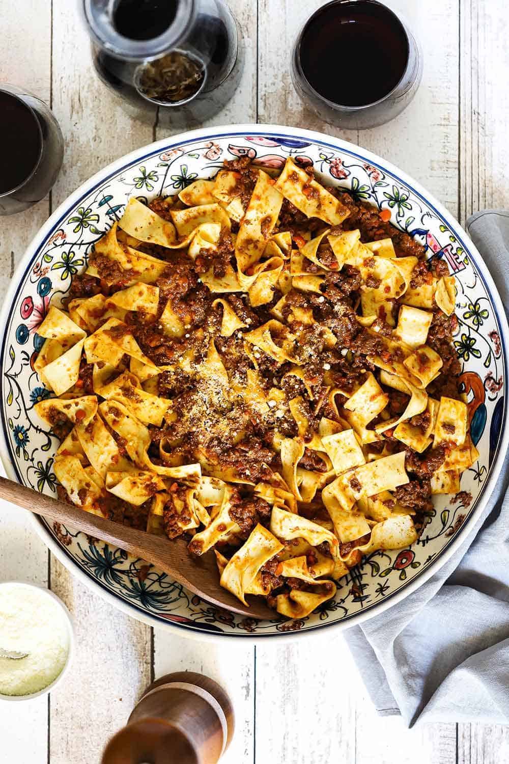 A large patterned Italian pasta bowl filled with Pappardelle Bolognese and a small bowl of Parmesan cheese next to it.