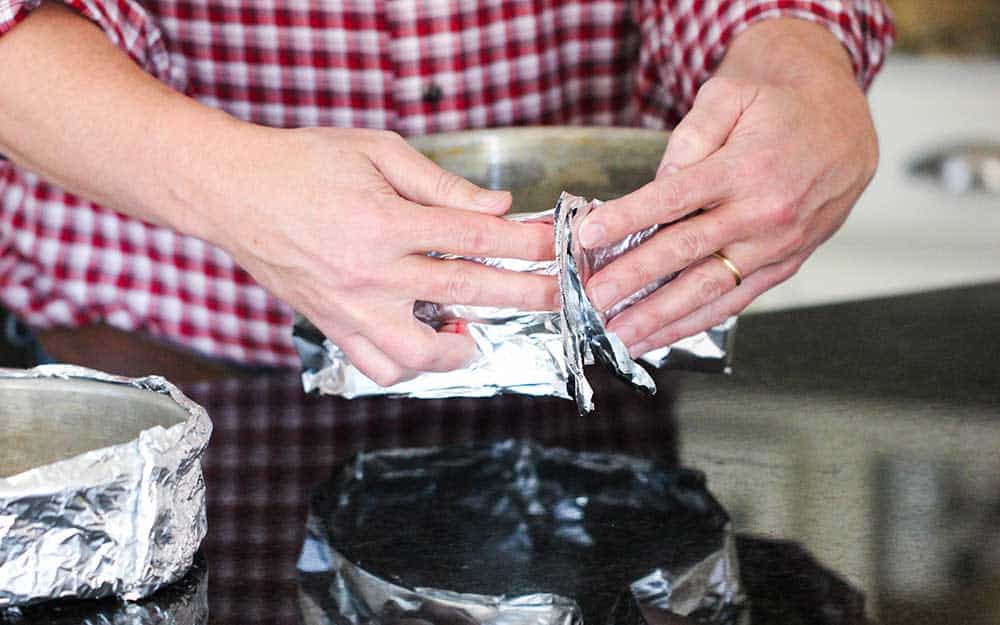 Two hands wrapping foil baking strips around a metal cake pan. 