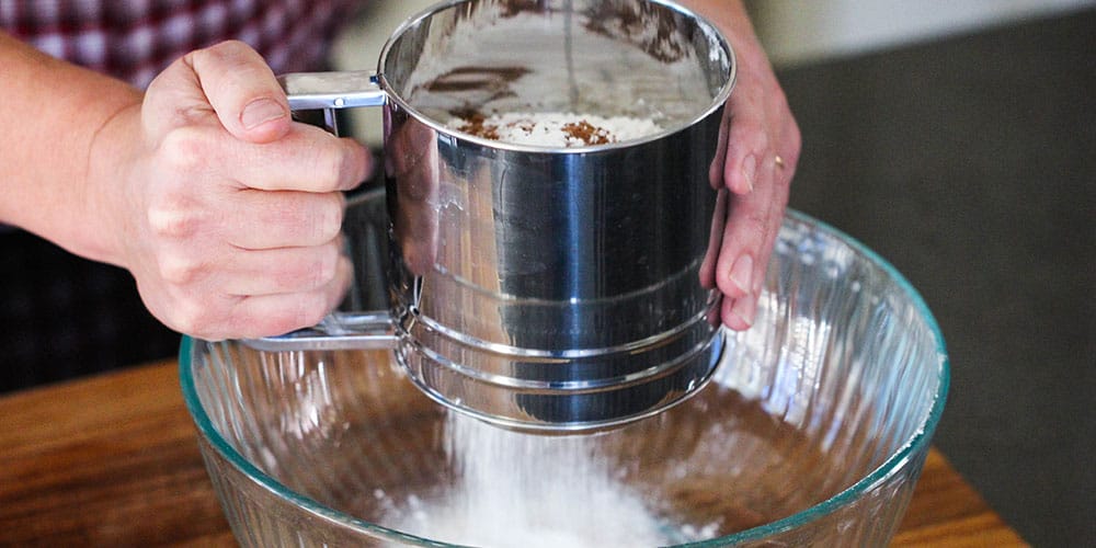 Two hands sifting flour and other dry ingredients into a glass bowl. 
