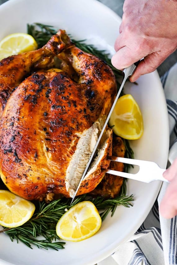 A person using a large fork and knife to slice in a roast chicken on a platter with cut lemons and herbs surrounding the bird.