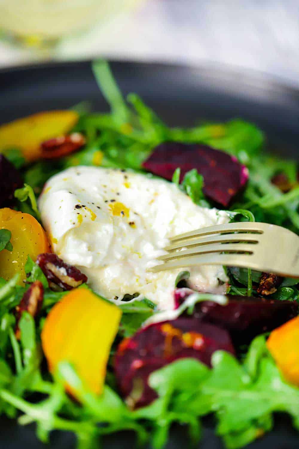 A fork breaking open a ball of burrata cheese sitting on top of sliced roasted beets and arugula salad. 