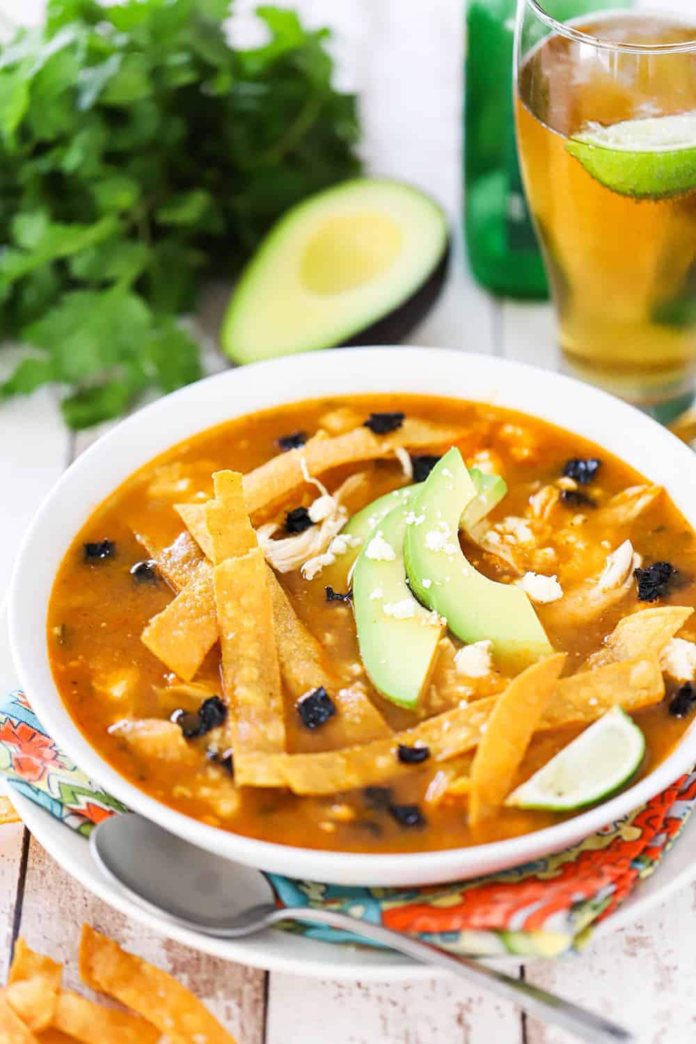 A white bowl filled with tortilla soup topped with avocado slices, tortilla strips, and a lime wedge all sitting next to a glass of beer. 