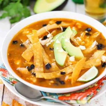 A bowl of chicken tortilla soup in a white bowl surround by a glass of beer and slice avocado.