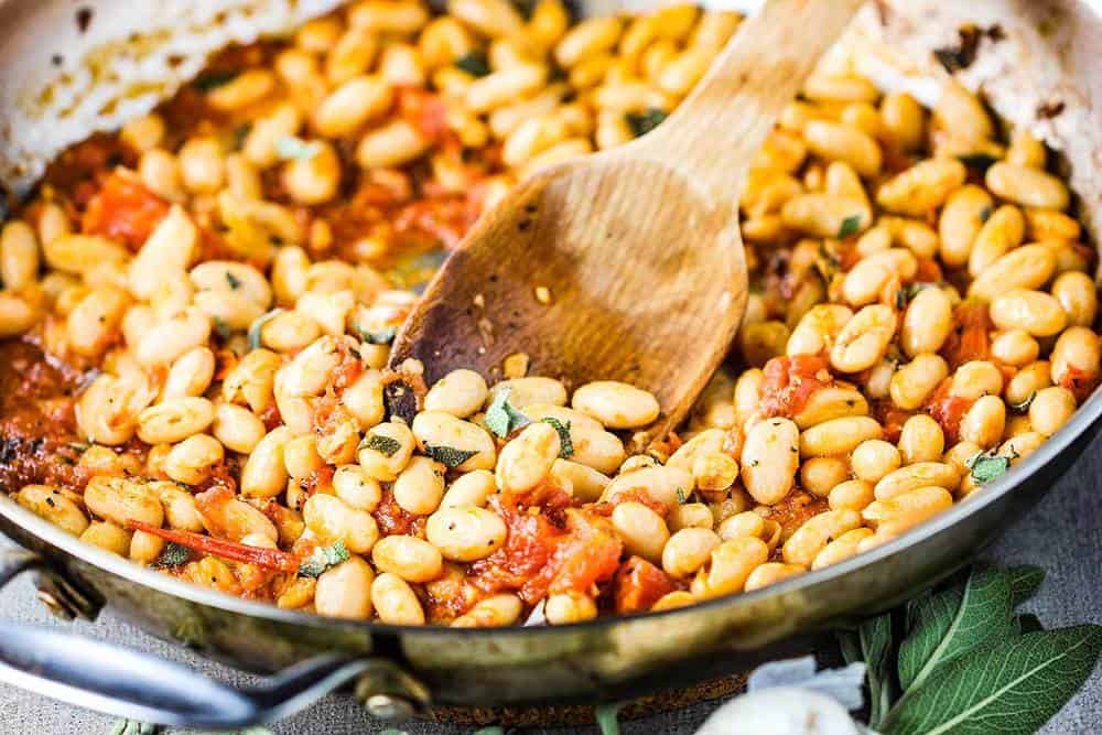 Sautéed white beans with garlic, sage and tomatoes in a skillet with a wooden spoon.