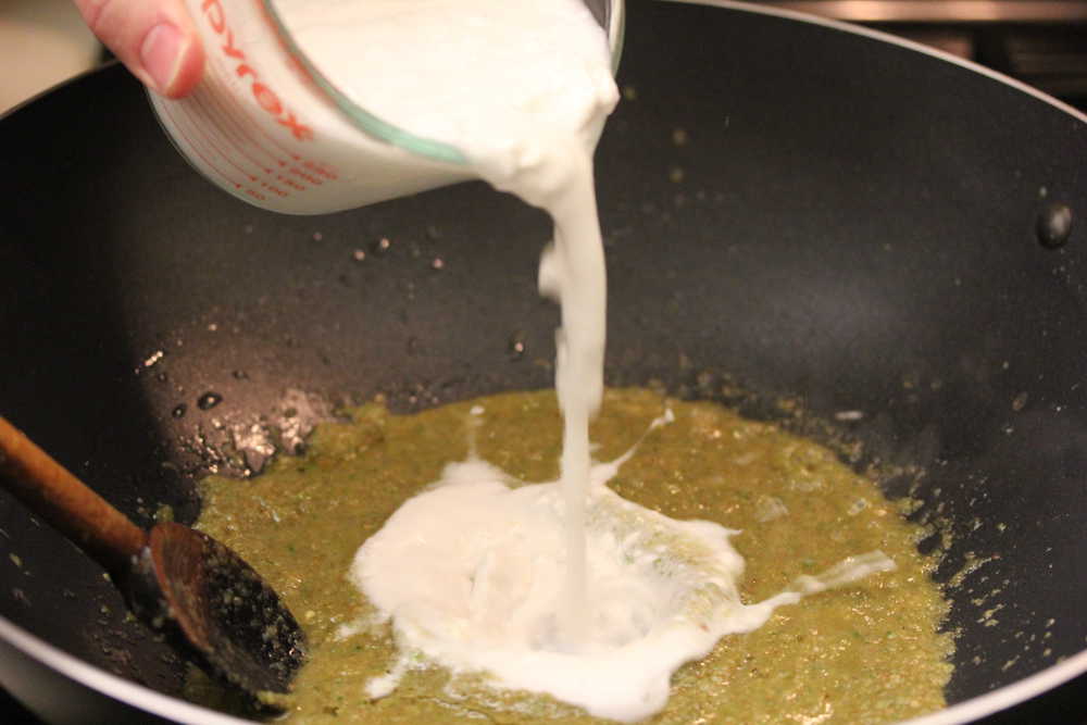 A glass measuring cup filled with coconut milk is being poured into a wok filled with green curry paste. 