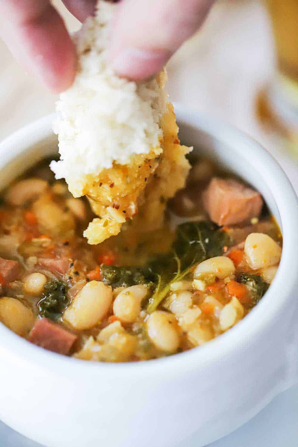 A hand dipping a torn piece of bread dipping into a bowl of smoked sausage and white bean stew. 