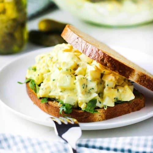 Best-ever egg salad sandwich on a white plate next to pickles